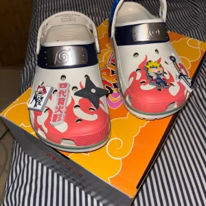 Naruto-inspirerade Crocs med dekorationer - Unika Crocs med Naruto-tema, dekorerade med karaktärer och symboler från serien. Skorna är vita med röda detaljer och har en blå rem. Perfekta för fans av anime och manga.