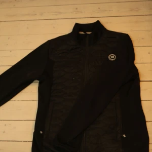 Pelle P cardigan - Snygg svart quiltad jacka från Pelle P med dragkedja framtill. Jackan har en stilren design med långärmade ärmar och en hög krage. Perfekt för kyligare dagar.