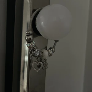 Silverfärgat armband med berlocker - Charmigt silverfärgat armband med flera berlocker, inklusive hjärtan och en blomma. Armbandet har glittriga och vita pärlor som ger en elegant touch. Perfekt för att lägga till lite bling till din outfit.
