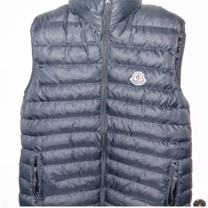 Svart dunväst från Moncler - Snygg svart dunväst från Moncler med dragkedja och hög krage. Perfekt för kyliga dagar. Västen har två praktiska fickor framtill och Monclers logga på bröstet.