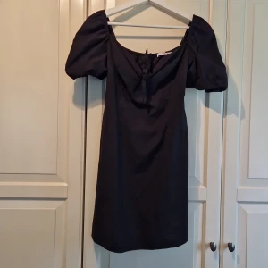 Svart klänning med puffärmar från NA-KD - Säljer en elegant svart klänning från NA-KD med puffiga kortärmar och en söt knytdetalj framtill. Klänningen har en smickrande passform och är perfekt för festliga tillfällen. Den är i nyskick och redo att bäras till nästa event!