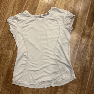 Vit Dri-FIT träningstopp från Nike - En vit Dri-FIT träningstopp från Nike. Den har en enkel och stilren design med korta ärmar och en liten Nike-logga på bröstet. Perfekt för träning eller en aktiv vardag! Storlek M