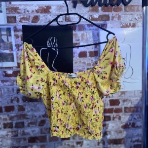 Gul blommig topp från FB Sister - Säljer en superfin gul topp från FB Sister med blommigt mönster i rosa och grönt. Toppen har korta ärmar med volangdetaljer och en smockad, croppad passform. Perfekt för sommardagar eller en festlig kväll!