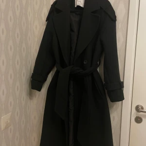 Svart Trench Coat Kappa  - Svart Vinter/höst kappa i modell av en trench coat i nyskick då den köptes under våren och är knappt använd från Bershka strl S.  Nypris 699kr 