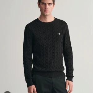 Svart kabelstickad tröja från Vivienne Westwood - Snygg svart kabelstickad tröja från Vivienne Westwood med ett diskret broderat emblem på bröstet. Tröjan har långa ärmar och ribbade muddar vid ärmslut och nederkant. Perfekt för en stilren look.