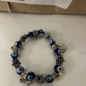 Blått armband med ögonmotiv och hamsa - Unikt armband med blå och silvriga pärlor, dekorerat med ögonmotiv och hamsa-symboler i silverfärg. Perfekt för att ge en bohemisk touch till din stil.