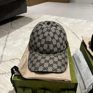 Gucci keps  - Säljer en snygg Gucci keps i beige med det klassiska GG-monogrammet i svart. Kepsen har en justerbar rem baktill och är i storlek M (58 cm). Perfekt för att ge din outfit en lyxig touch! 🧢 