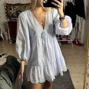 Ljusblå randig klänning med broderi - Superfin ljusblå och vit randig klänning med v-ringning och volanger. Den har långa puffärmar med vackert broderi och knytning framtill. Perfekt för vår och sommar!