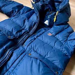 Blå dunjacka från Fjällräven - Säljer en snygg blå dunjacka från Fjällräven, perfekt för kalla vinterdagar. Jackan har en praktisk huva med gula detaljer och dragkedja framtill. Två stora fickor med knappar ger extra förvaring. Den är i mycket bra skick och redo för nya äventyr! 💙