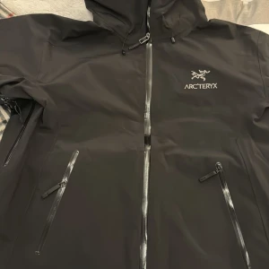 Svart regnjacka från Arc'teryx - Säljer en svart regnjacka från Arc'teryx i mycket bra skick. Jackan har en justerbar huva och dragkedja framtill. Perfekt för regniga dagar och utomhusaktiviteter. Den är både stilren och funktionell.