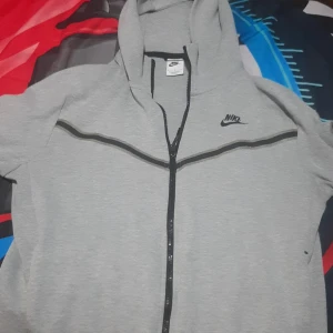 Grå Nike tech - Säljer en grå hoodie från Nike i bra skick. Den har en dragkedja framtill och en klassisk Nike-logga på bröstet. Perfekt för träning eller en avslappnad dag. Den är bekväm och stilren med svarta detaljer längs dragkedjan och ärmarna.