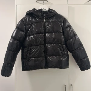 Svart pufferjacka med huva - Snygg svart pufferjacka med glansig yta och huva. Jackan har en dragkedja framtill och långa ärmar. Perfekt för kyliga dagar.