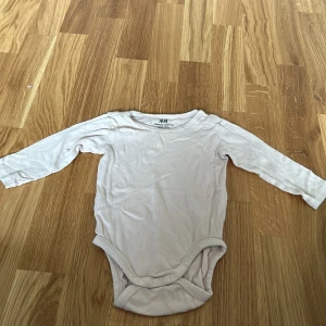 Vit långärmad body från H&M - Säljer en söt och bekväm vit långärmad body från H&M i ekologisk bomull. Perfekt för de små under vår och höst. Praktiska knappar i grenen för enkel på- och avklädning.