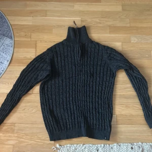 Mörkgrå stickad tröja från Indicode - Säljer en mörkgrå stickad tröja från Indicode med dragkedja och hög krage. Tröjan har ett ribbat mönster och långa ärmar, perfekt för kyliga dagar. Passar bra till både jeans och chinos.