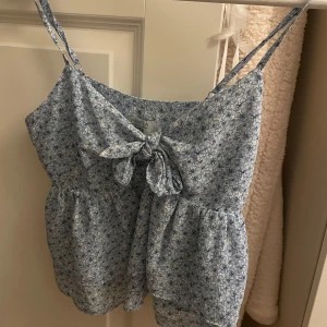 Blommig topp från NA-KD - Söt blommig topp från NA-KD i ljusblått med smala axelband och knytning framtill. Band som går att justera och knappt aldrig använd🩷