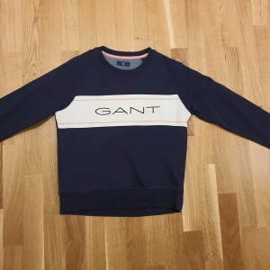 Mörkblå sweatshirt från GANT - Säljer en snygg mörkblå sweatshirt från GANT med en vit och rosa rand över bröstet och GANT-loggan i svart. Tröjan har långa ärmar och en rund halsringning med ribbade kanter. Perfekt för en avslappnad stil.