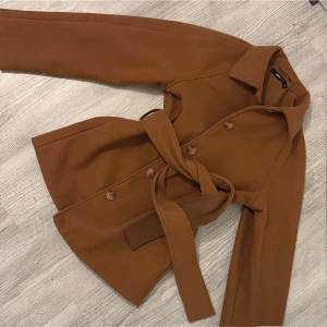 Brun kappa med bälte - Snygg brun kappa med knappar och bälte i midjan. Perfekt för höst och vår. Långärmad design med klassisk krage. Funkar bra till vintern med! Använd en gång nypris 600