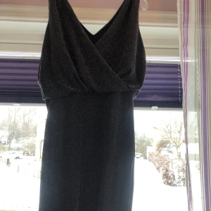 Svart glittrig klänning - Elegant svart klänning med glittrigt mönster och omlott-design. Den har en v-ringad hals och är ärmlös, vilket ger en stilren look. Perfekt för festliga tillfällen.