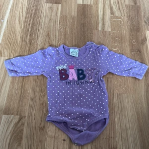Lila prickig babybody - Söt lila babybody med vita prickar och texten 'New Baby in Town' i olika färger och mönster. Bodyn har långa ärmar och praktiska knappar vid axeln för enkel påklädning. Perfekt för de små! 💜