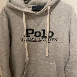 Grå hoodie från Polo Ralph Lauren - Säljer en klassisk grå hoodie från Polo Ralph Lauren med logotyp framtill. Den har en justerbar huva med snören och en stor magficka. Perfekt för en avslappnad stil.