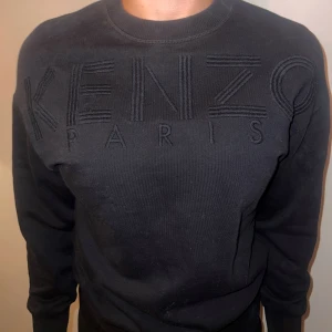 Svart tröja från Kenzo - Säljer en stilren svart tröja från Kenzo med deras ikoniska logga broderad på framsidan. Tröjan har långa ärmar, breda muddar och en normal passform, perfekt för både vardag och fest. Den är i mycket bra skick och redo för en ny ägare!