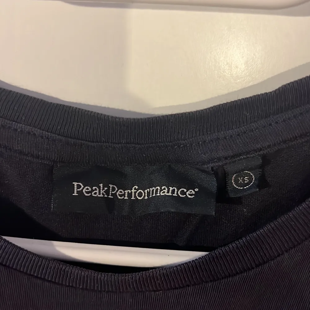 Säljer en svart/mörkblå t-shirt från Peak Performance i storlek XS. Den har ett coolt tryck med texten 'Peak Performance' i vitt på framsidan. Perfekt för en avslappnad stil eller träning. T-shirten är i bra skick och gjord av mjukt material. Knappast använd.. T-shirts.