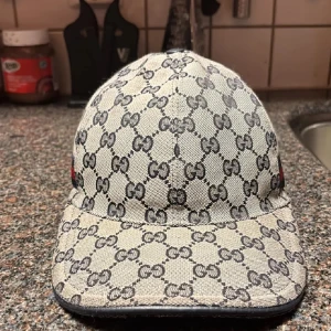 Gucci keps  - Snygg keps från Gucci med det ikoniska GG-mönstret i grått och svart. Kepsen har en justerbar rem baktill och en röd och blå randdetalj på sidan. Perfekt för att ge din outfit en lyxig touch.