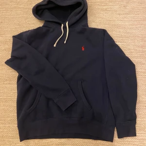 Blå hoodie från Ralph Lauren - Säljer en klassisk blå hoodie från Ralph Lauren med den röda logotypen på bröstet. Tröjan har en justerbar huva med vita snören och en praktisk magficka. Perfekt för hösten 