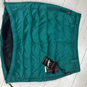 Grön quiltad kjol från Skhoop - Snygg och praktisk grön quiltad kjol från Skhoop. Perfekt för kyliga dagar med sin vadderade design och dragkedjor på sidorna för enkel på- och avtagning. Kjolen har en sportig look och är idealisk för utomhusaktiviteter.