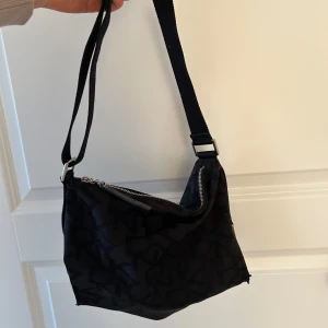 Svart axelväska från DKNY - Snygg svart axelväska från DKNY med ett diskret mönster och dragkedja upptill. Väskan har en justerbar axelrem och är i mycket bra skick.