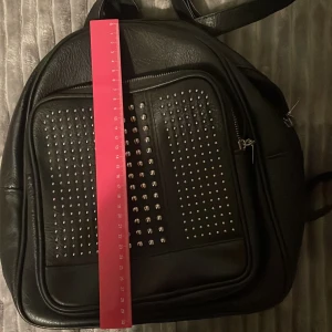 Säljer en fin svart väska med unika detaljer - Väskan är ungefär 30-31 cm lång och ca 13 cm bred. Det väskan är svart och har unika silver detaljer på ytterfacket. Den är i hyfsat nyskick utan skador. 