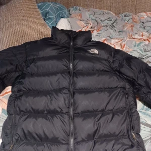 Svart dunjacka från The North Face - Säljer en svart dunjacka från The North Face. Jackan har en dragkedja framtill och är långärmad. Perfekt för kalla dagar med sin pufferdesign som håller värmen.