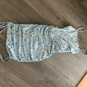 Blommig blå klänning med knytband - Söt blå klänning med blommigt mönster och smala axelband. Klänningen har knytband på sidorna för justerbar passform och en rynkad effekt. Perfekt för sommarens varma dagar.