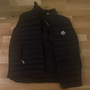 Svart dunväst Moncler - Säljer en stilren svart dunväst Moncler med dragkedja och broderad logga på bröstet. Perfekt för kyligare dagar och enkel att matcha med olika outfits.