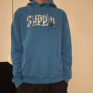 Blå hoodie från Supply & Demand - Säljer en blå hoodie från Supply & Demand med tryck framtill. Den har en klassisk känguruficka och justerbar huva med dragsko. Perfekt för en avslappnad stil.