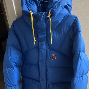 Blå dunjacka från Fjällräven - Säljer en blå dunjacka från Fjällrävens 1974 Expedition Series. Jackan har en huva med gula snören och knäppning med både dragkedja och knappar. Den är nytvättad det är därför den är i en påse. Så den e som ny