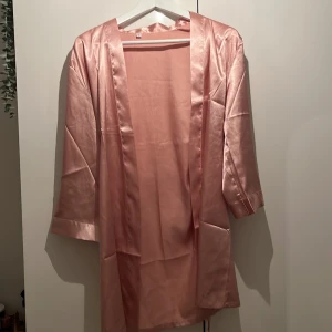 Rosa sidenkimono - Elegant rosa kimono i siden med trekvartsärm. Perfekt för en lyxig känsla hemma eller som ett stilrent lager över en outfit. Den har en mjuk och glansig yta som ger en exklusiv look. Vet inte märket då jag fick den för längesedan