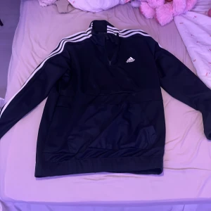 Svart tröja från Adidas - Säljer en svart tröja från Adidas med klassiska vita ränder längs ärmarna och en halv dragkedja framtill. Perfekt för en sportig look eller avslappnad stil. Adidas-logga på bröstet.