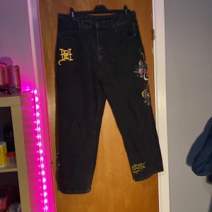 Ed Hardy jeans - Snygga svarta jeans från Ed Hardy med ett coolt drakbroderi i lila, rött och gult på benet. Byxorna har en normal passform och är dekorerade med Ed Hardy-logga i gult på baksidan. Perfekta för dig som vill sticka ut!