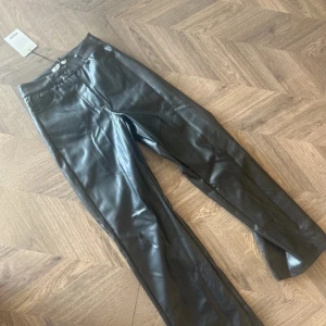 Svarta skinnbyxor från NA-KD - Snygga svarta skinnbyxor från NA-KD med hög midja och knäppning fram. Perfekta för en trendig look med en tight passform som framhäver figuren. Byxorna har en klassisk design med fickor och bälteshällor.