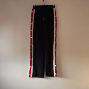 Svarta byxor med knappar - Snygga svarta byxor med röda och vita detaljer längs sidorna. Byxorna har knappar hela vägen ner för en cool look. Perfekta för en avslappnad stil med en twist.