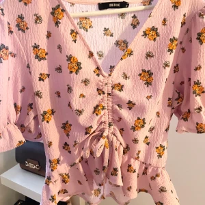 Rosa blommig blus från Bik Bok - Söt rosa blus med blommigt mönster i orange och grönt från Bik Bok. Blusen har en v-ringad design med knytning framtill och korta puffärmar. Perfekt för en somrig look!