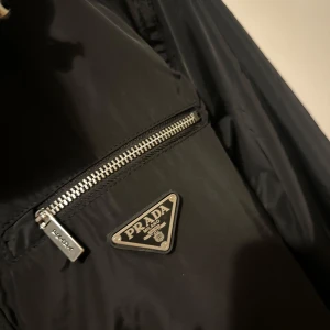 Svart jacka från Prada - Snygg svart jacka från Prada med dragkedja och knappar. Jackan har en praktisk huva och en bröstficka med dragkedja. Perfekt för en stilren look. 