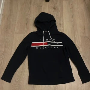 Marinblå hoodie från Tommy Hilfiger - Snygg marinblå hoodie från Tommy Hilfiger med klassisk logga i rött, vitt och blått på bröstet. Tröjan har en justerbar huva med vita snören och långa ärmar. Perfekt för en avslappnad stil. 8/10
