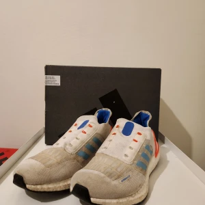 Adidas Ultraboost i vitt och blått - Säljer ett par Adidas Ultraboost sneakers i vitt med blå och orange detaljer. Skorna har en bekväm Boost-sula och är perfekta för löpning eller vardagsbruk. De har en stickad ovandel för extra komfort. Skorna har 41.5 som storlek men passar 40-42. 