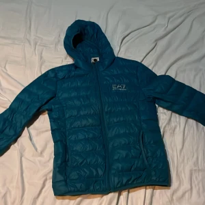 Blå dunjacka från Emporio Armani - Snygg blå dunjacka från Emporio Armani EA7 med huva och dragkedja. Perfekt för kyliga dagar med sin stilrena design och bekväma passform.