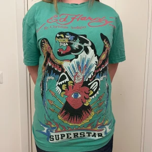 Grön t-shirt från Ed Hardy - Cool grön t-shirt från Ed Hardy med ett stort, färgglatt tryck av en tiger och texten 'SUPERSTAR' på framsidan. T-shirten har korta ärmar och är i en normal passform. Perfekt för dig som vill vara SKIT BALL😸 As skön tröja som är lite längre!💞