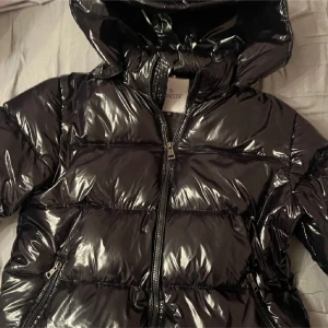 Svart pufferjacka från Moncler - Snygg svart pufferjacka från Moncler med glansig finish och huva. Jackan har en dragkedja framtill och Moncler-logga på ärmen. Perfekt för kyliga dagar. Pris går att diskutera 