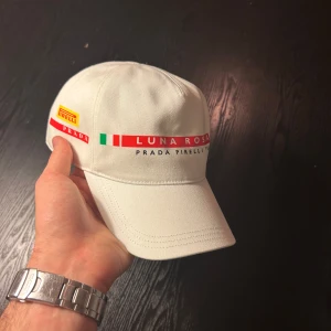 Vit keps från Prada - Snygg vit keps från Prada x Pirelli med texten 'Luna Rossa' och italienska flaggan på framsidan. Kepsen har en klassisk design med böjd skärm och justerbar passform. Perfekt för en stilren look.