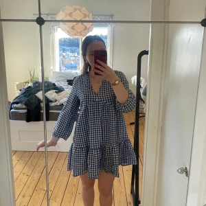 Blårutig klänning med volang - Charmig blårutig klänning med trekvartsärm och volangdetalj. Perfekt för en avslappnad stil med en luftig passform. Den har en v-ringad hals och är gjord i ett lätt material som ger en bekväm känsla.